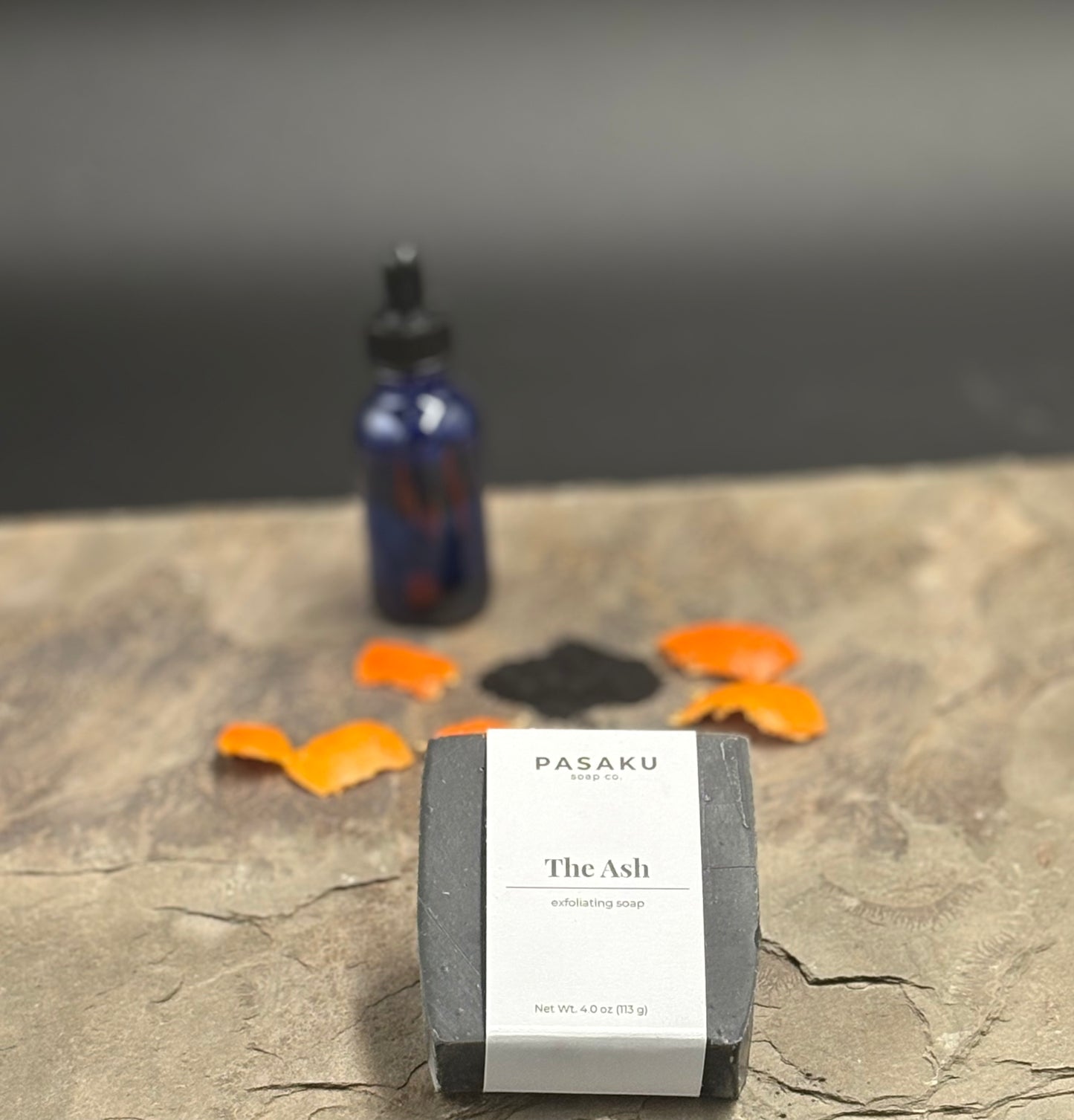 Black bar of exfoliating soap with 'Pasaku Soap Co' on the label on a slate and gray background. Included on the label are the words 'The Ash' and 'exfoliating soap'. In the background is a blurred bottle, orange peel and a pile of charcoal.