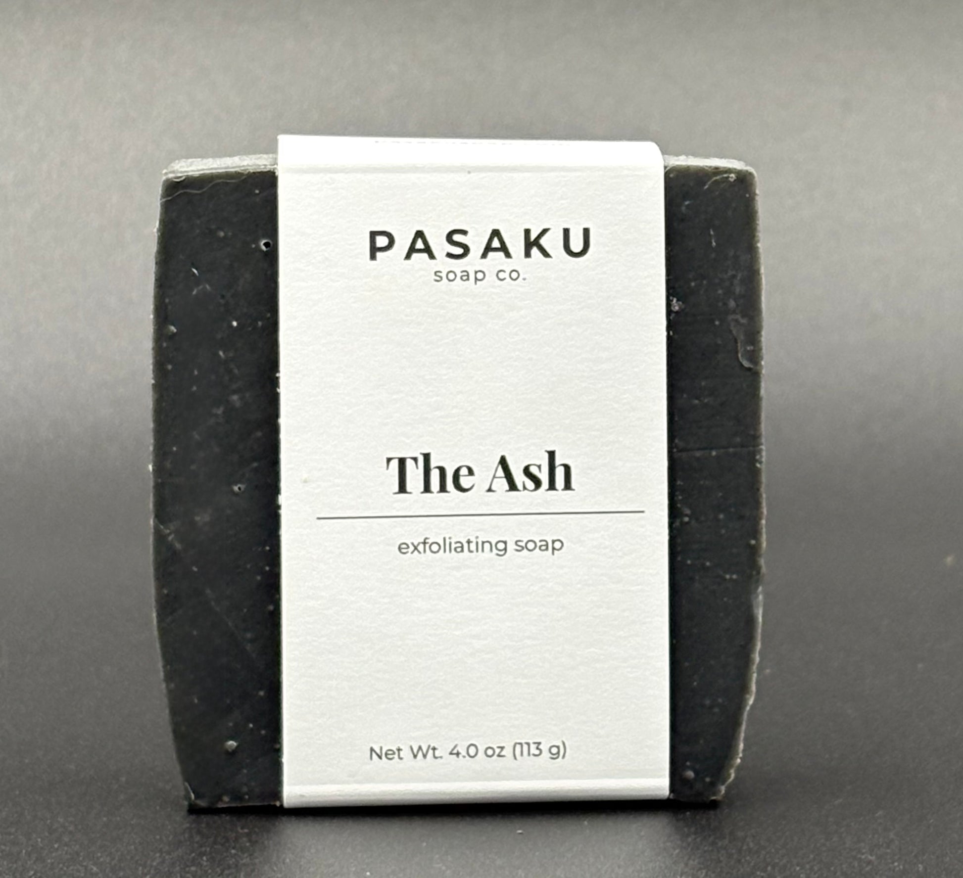 Black bar of exfoliating soap with 'Pasaku Soap Co' on the label on a gray background.  Included on the label are the words 'The Ash' and 'exfoliating soap'.