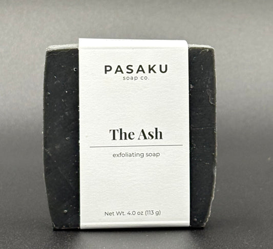 Black bar of exfoliating soap with 'Pasaku Soap Co' on the label on a gray background.  Included on the label are the words 'The Ash' and 'exfoliating soap'.