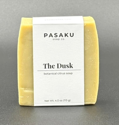 A cream-colored soap bar, on a blank background.  The label wrapped around the bar has the name of the company, Pasaku Soap Co., with the name of the soap, The Dusk, and the words botanical citrus soap.