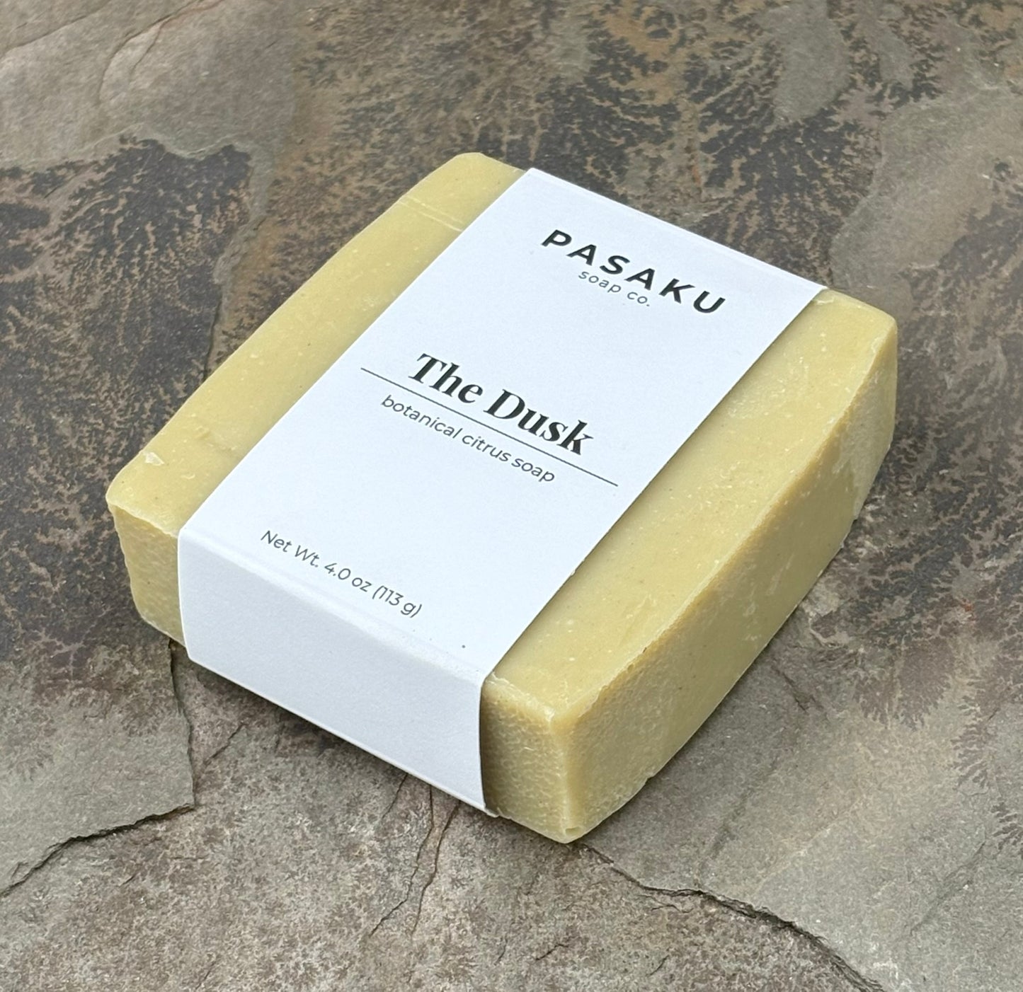 A cream-colored soap bar, on a piece of slate.  The label wrapped around the bar has the name of the company, Pasaku Soap Co., with the name of the soap, The Dusk, and the words botanical citrus soap.