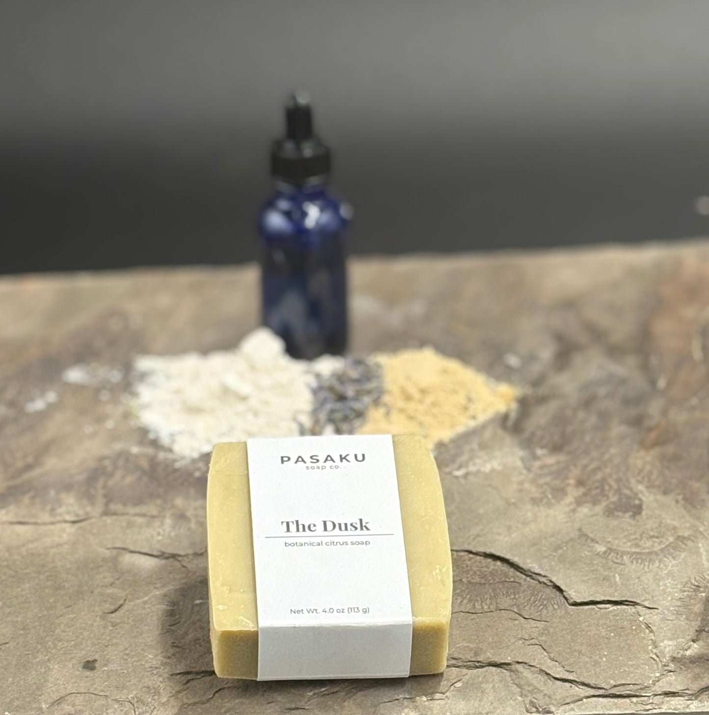 A cream-colored soap bar, on a piece of slate.  The label wrapped around the bar has the name of the company, Pasaku Soap Co., with the name of the soap, The Dusk, and the words botanical citrus soap.  In the background is an apothecary bottle, with piles of clays and lavender in the background.