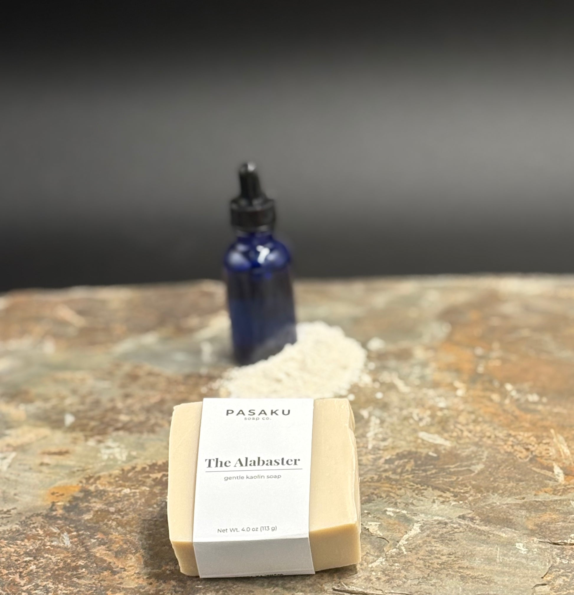 with white wrap around label that has the name of the company, the name of the soap, and the net weight of 4 oz. set on a slate surface, with a blue apothecary jar in the background with a pile of kaolin clay.